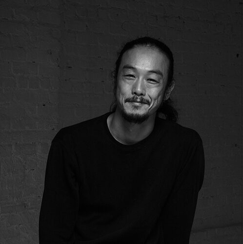 A black and white image of a Japanese man with hair tie back, smiling and seating on a high stool.