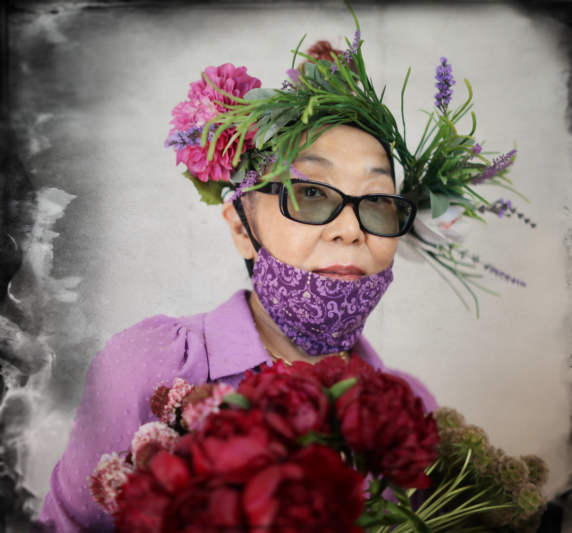 Joan Osato is in a purple shirt with a purple mask. She wears black framed glasses and carries flowers.