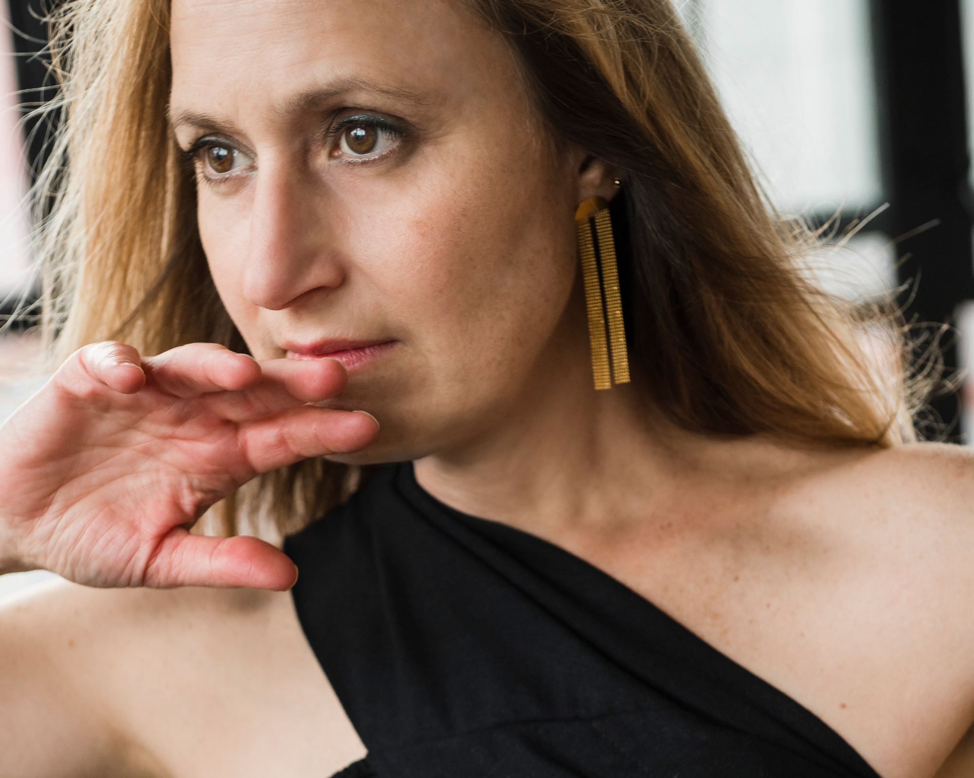 A blonde, light skinned Middle Eastern American woman looking far into the distance with her hand delicately at her mouth as if asking a question.