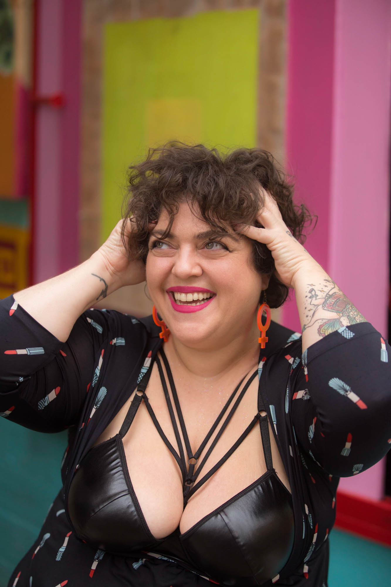 An Arab-American woman with short curly hair, wearing venus earrings, a bustier, and a black and white faux fur collar adjusts an invisible crown on her head, joyfully