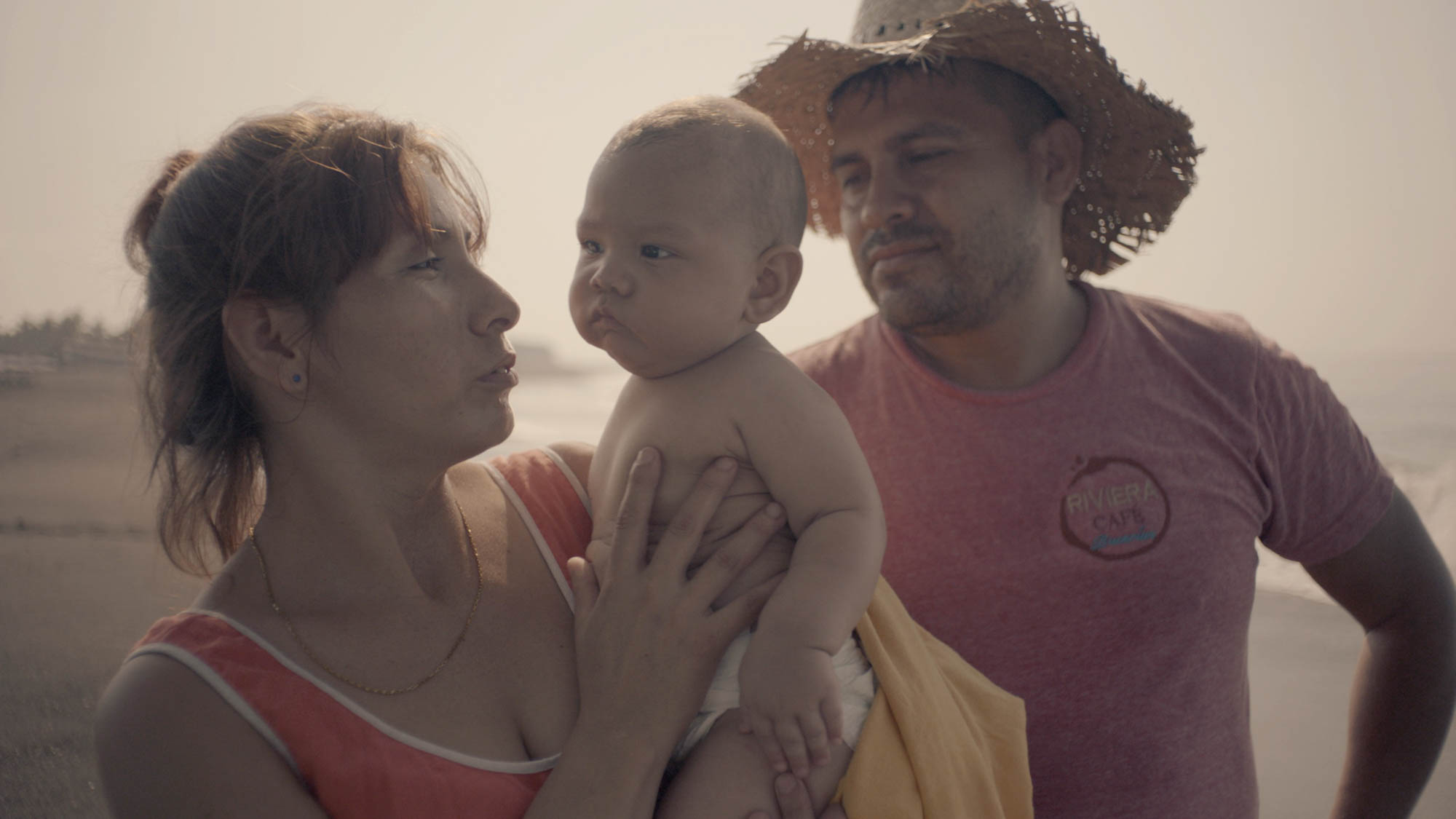 A couple stands on a beach awash in sun. The woman is in the foreground, holding up a beautiful infant child. The father stands close but a little further back, wearing a straw hat and watching both his wife and his son.