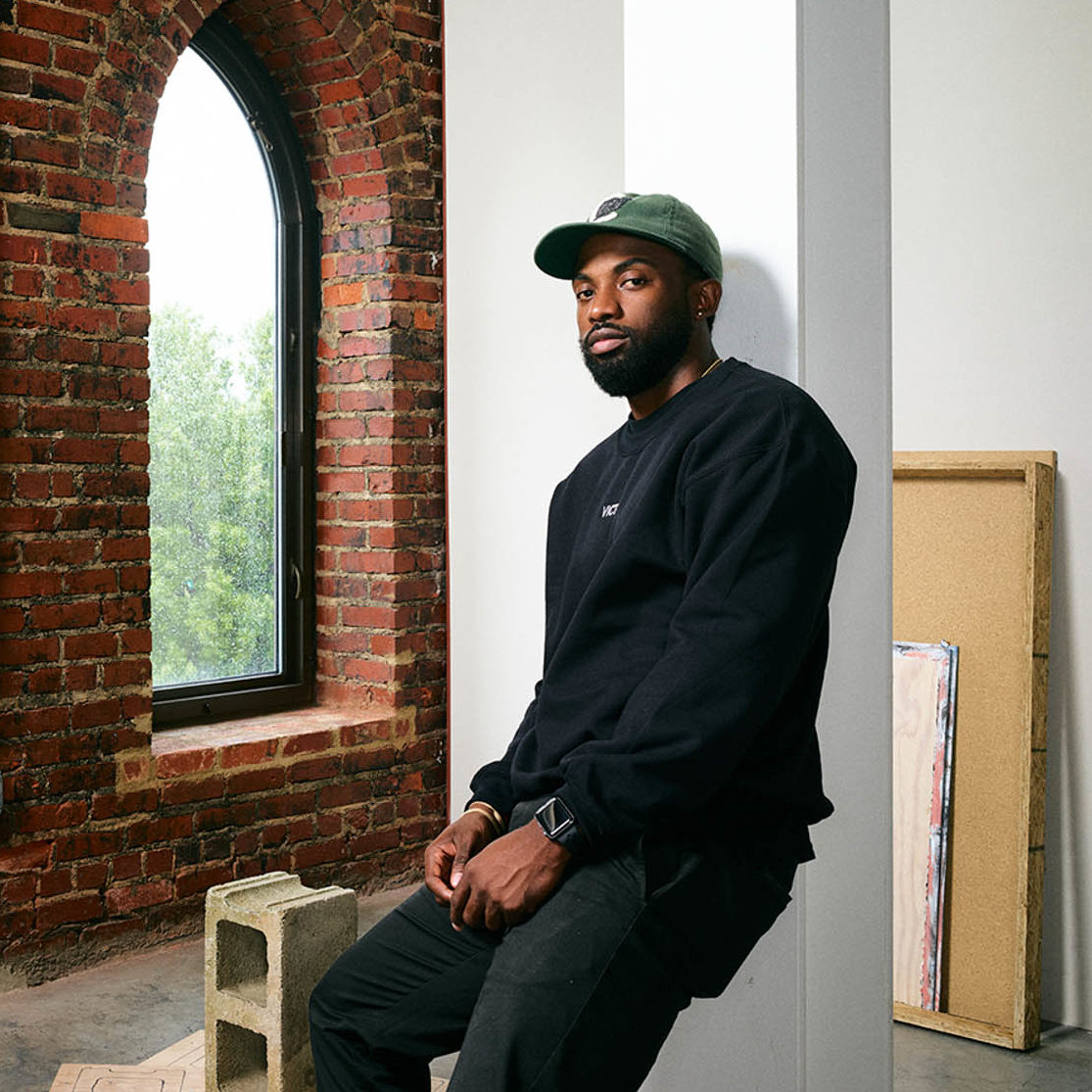 An African American man, wearing green baseball cap, black sweatshirt and black pants leaning on white steel I-beam pillar. Behind him is the corner of a studio, with the left wall expose brick and the perpendicular right wall painted white. A cinder block, plywood and wooden frames are laid on the floor and along the wall.