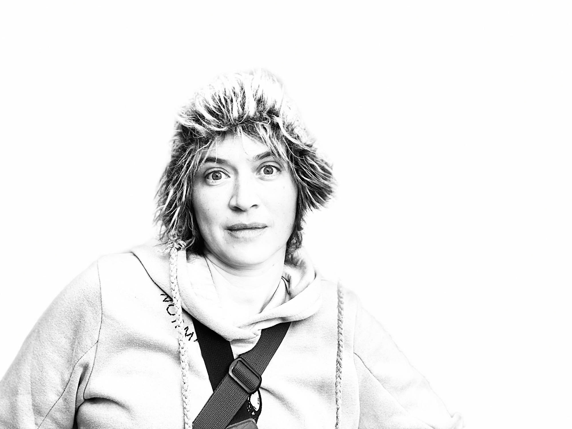 SUE-C (Sue Slagle) with fuzzy hat, hooded sweatshirt, and cameras strapped around her shoulder faces the viewer with large eyes.
