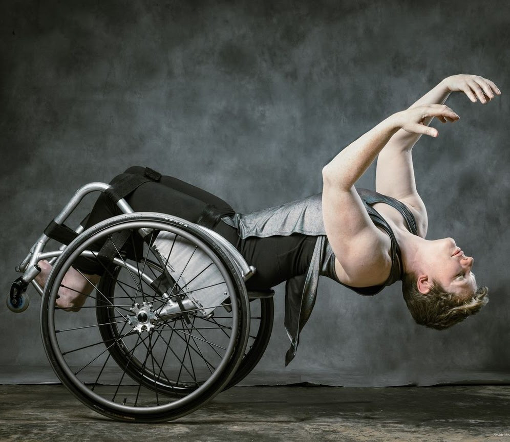 Laurel arches backward, balancing on two wheels, impossibly close to the floor. Arms curve loosely up over body and face; her silver top drapes and flows into her silver chair frame. Photo David Clifton-Strawn