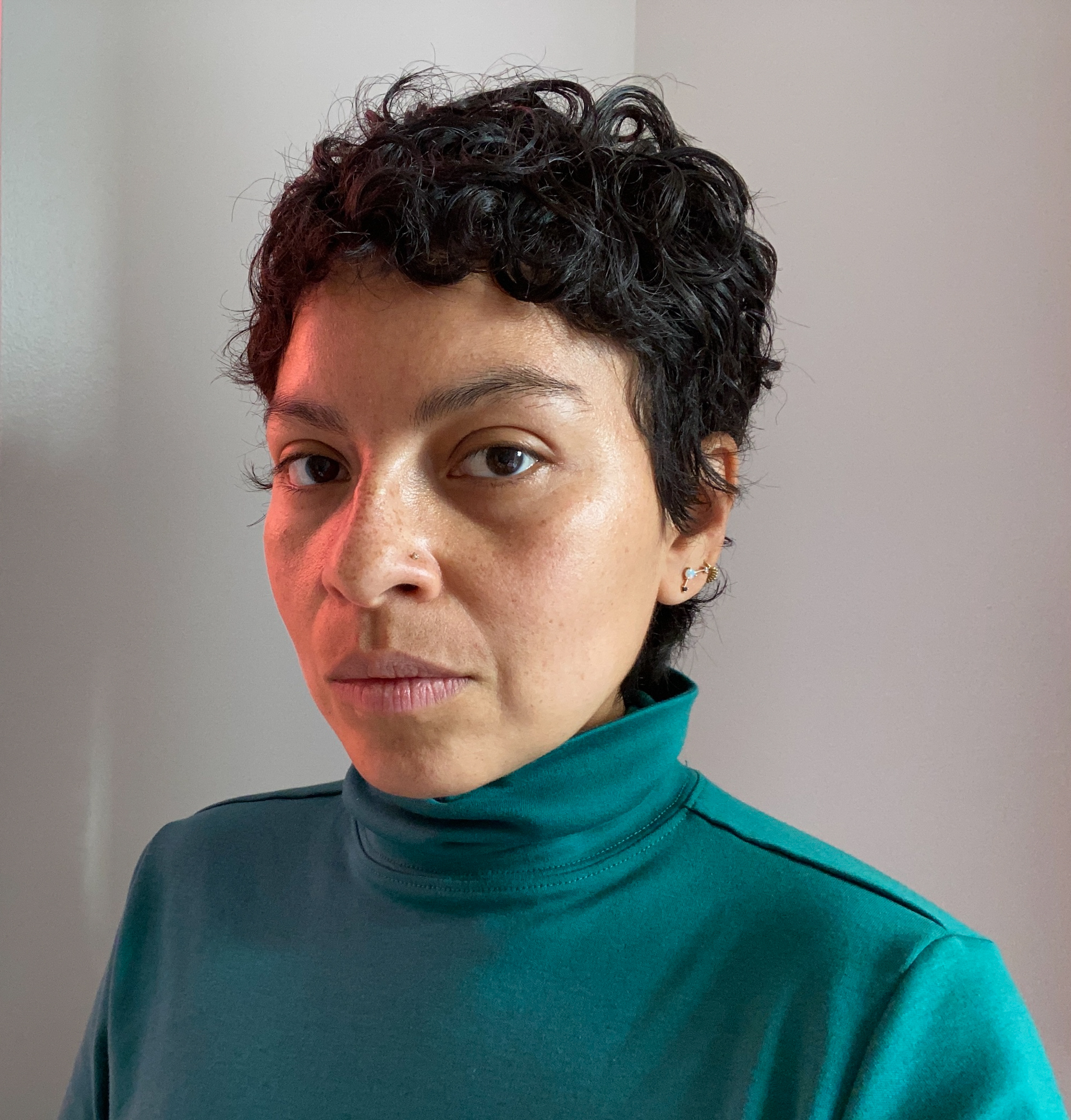 A Latinx woman with short black curly hair and tan skin looks straight into the camera, she wears a teal turtleneck.