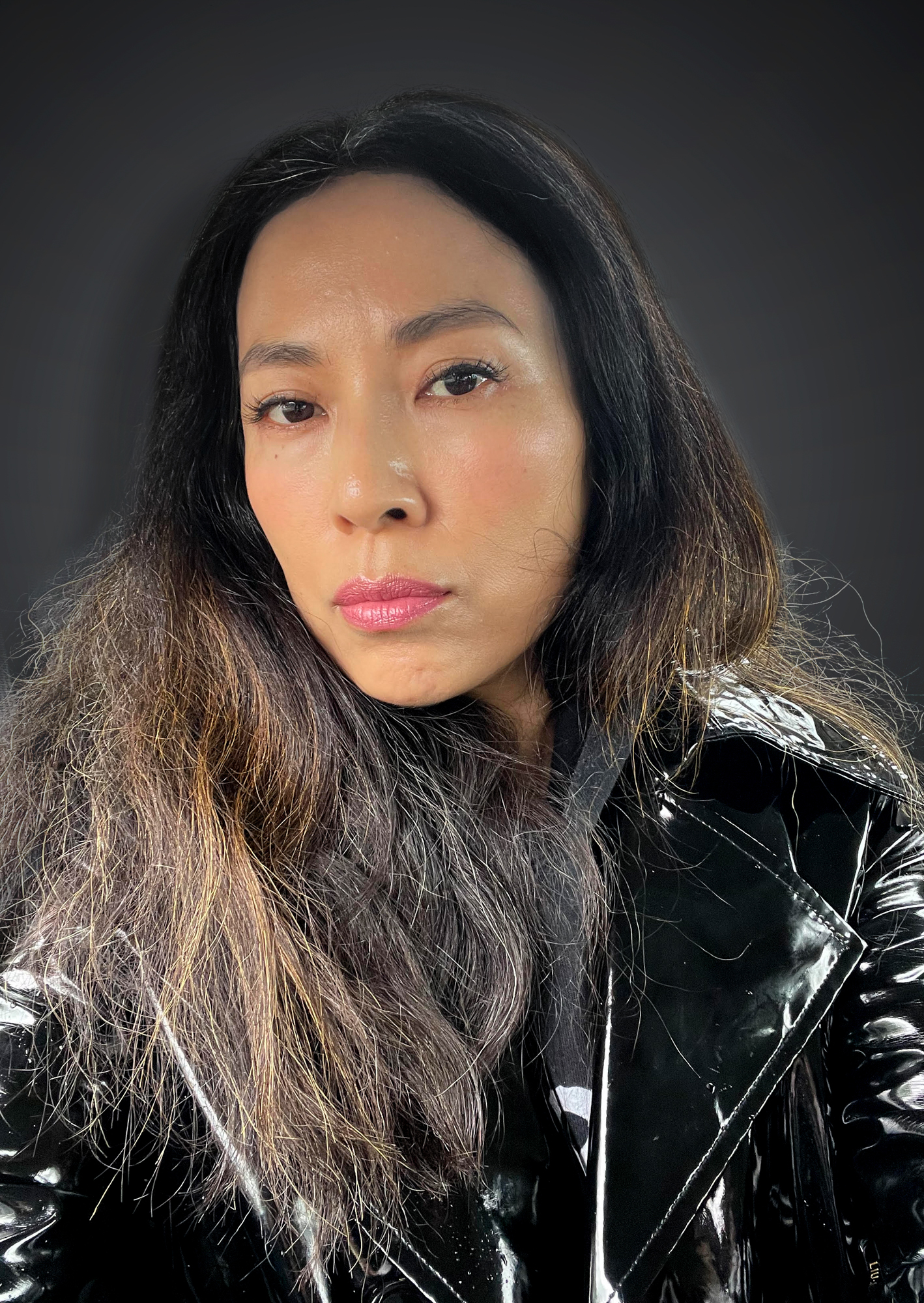 Headshot of artist Anicka Yi with long hair and wearing a shiny black trench coat and grey hoodie.