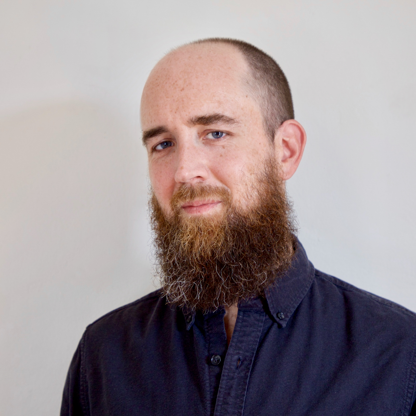 A man with a reddish beard, a high forehead, blue eyes, and freckles wearing a dark shirt with buttons is looking toward the camera with his head turned slightly.