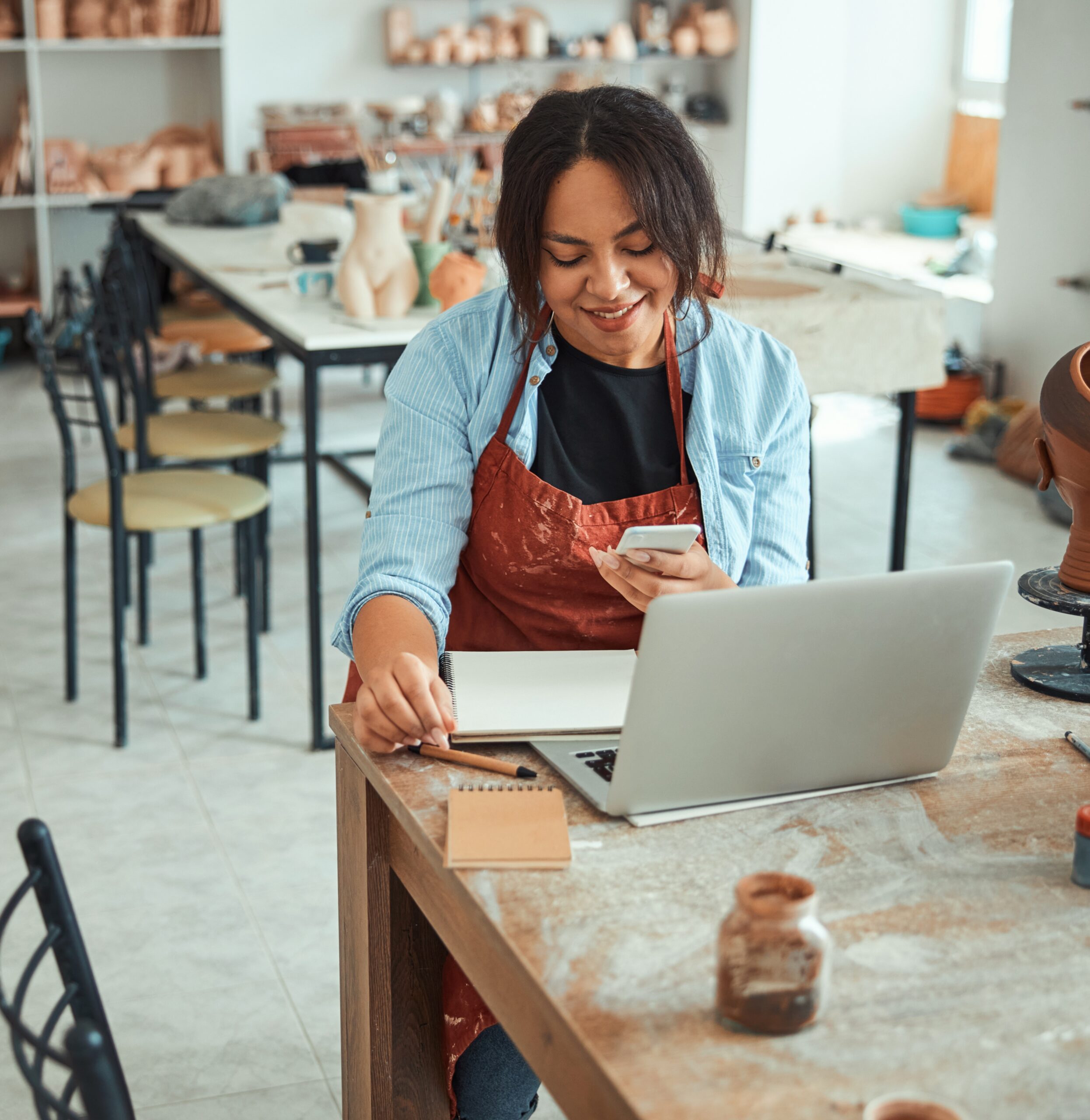 Charming female potter holding mobile phone and smiling while sitting at the table with notebook