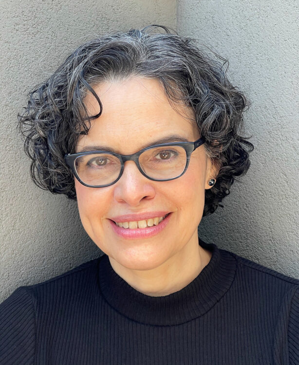 A woman with glasses, graying short, curly black hair, wearing a black mock turtleneck, in front of a gray wall.