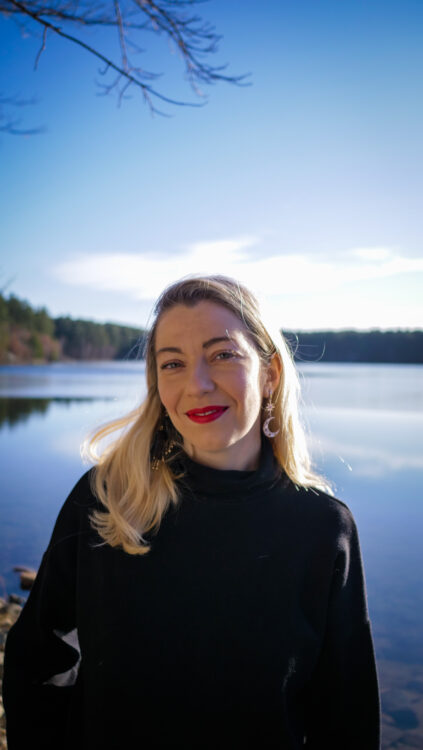 A caucasian woman with long blond hair, wearing a black sweater. Behind her is a lake, clear sky, and distant trees.