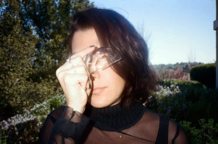 An analogue image of a woman with brown hair, holding an eye-shaped optical lens in front of her eye. Behind her is greenery and it's daylight.