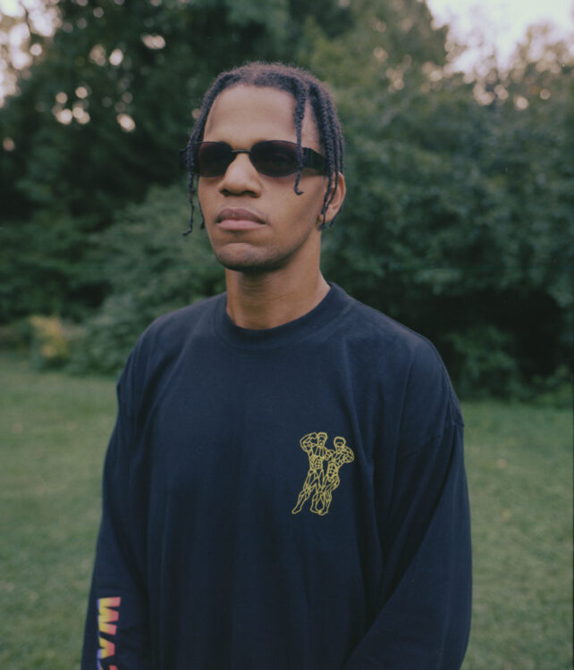 A portrait photo of a person from the midriff upwards. They have brown skin and braids that hang down beside their face to the cheek. They have dark sunglasses and no expression. They are wearing a baggy black long sleeve shirt with colorful print on their right sleeve and a small yellow figure on their left chest. They are standing in green grass with green trees behind them, the grass and trees nearly fill the remainder of the frame. The lighting is low as if it is early morning or late afternoon.
