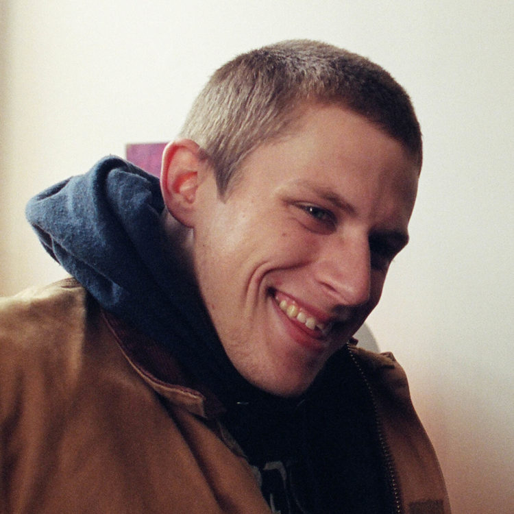 A young white man with a chipped tooth and short hair smiles in front of a white wall.