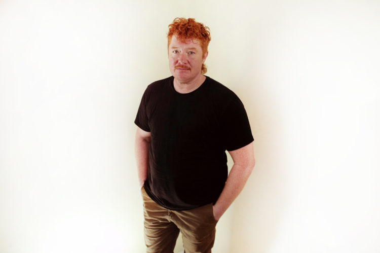 A masculine gender queer person standing with short orange hair smirking towards the camera. 