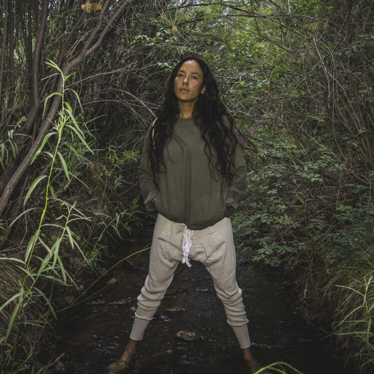 Shayla Blatchford, an Indigenous woman with long dark hair, stands in the middle of a serene stream surrounded by lush greenery. She wears an olive green jacket and looks directly at the camera with a calm and confident expression.