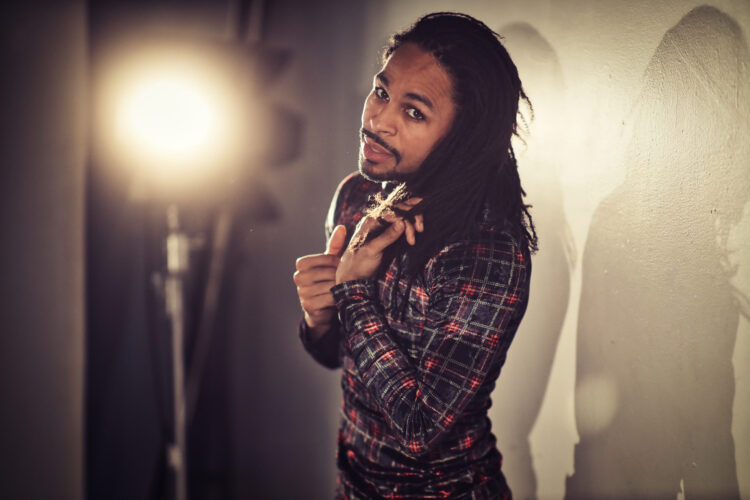 A non-binary artist with caramel skin, brown hazel eyes with thick, dark eyelashes runs their fingers through their shoulder length dark brown locs. They are wearing a plaid red, black and gold velvet turtleneck and stand in front of a stage light. Photo by Marcus Shields, 2021