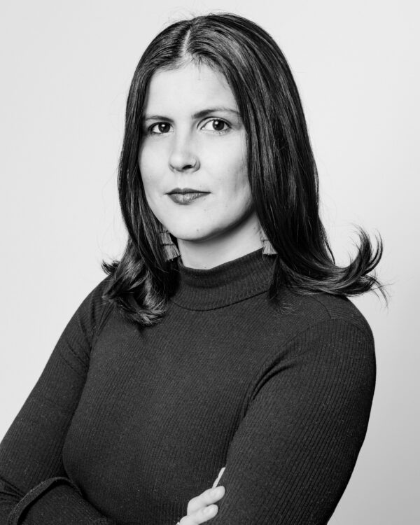 Black and white portrait of a light skinned female with shoulder length brown hair, black turtle neck sweater and fringe earrings.
