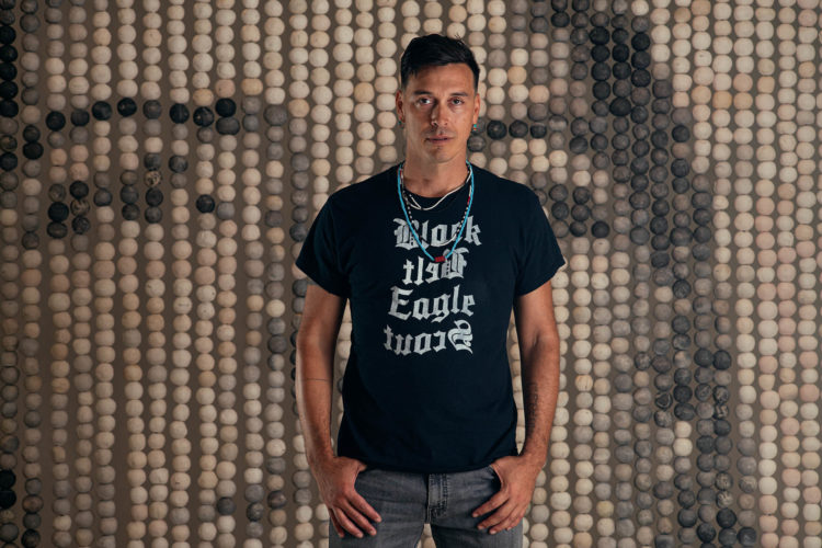 Artist Cannupa Hanska Luger wears a black shirt and beaded necklace, with hands in his grey jeans pockets, he is standing in front of his monumental clay bead artwork titled Every One. Photo: Brendan George Ko.