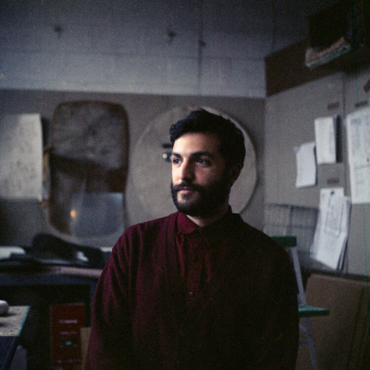 A bearded young Iranian man in maroon shirt looks off into the distance with faint smile, center framed in a warehouse studio, murky grey Chicago light diffused by dirty windows, shot on grainy expired film