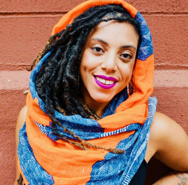 A bronze-skinned woman with fuscia lipstick, long locks, orange scarf framing her face smiles, exuding warmth, light, and renewal. 