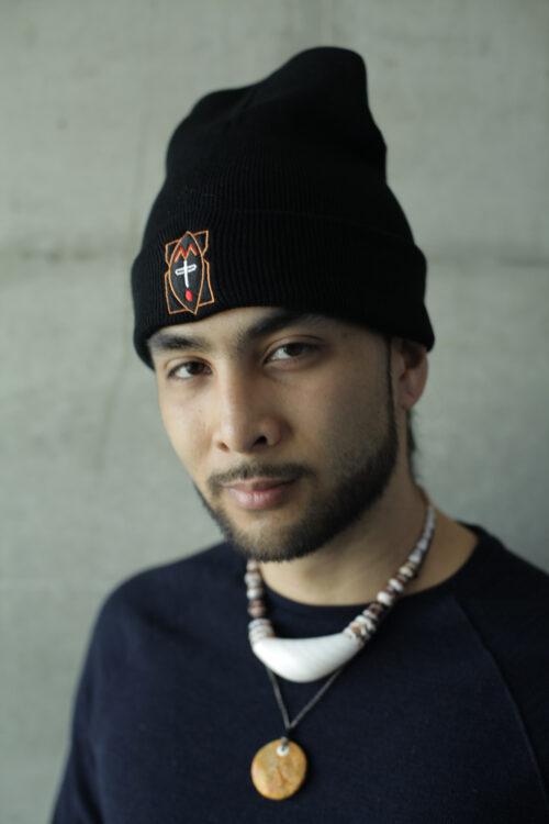 A brown-skinned Matao person looks curiously into the lens of a camera. The background is a grey wall and their image pops out the most. Their face is lined by very neatly cut facial hair and a black beanie with an ancestral symbol on it. Guiya (they) wear a sinahi and a tigem pendant.