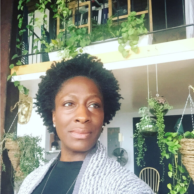 Black woman with a crown of kinky coily hair wearing a black shirt and grey sweater, and standing in front of a lush indoor garden.