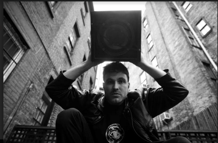This is the headshot of Devin B Waldman holding a speaker above his head. The photo is taken by Isaac Rosenthal in New York City in 2020.