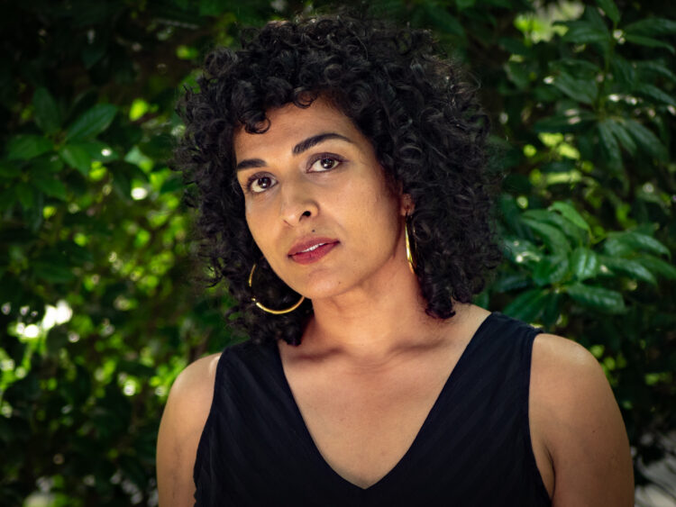 South Asian woman with black curly hair wearing a black jumpsuit and standing against a green leafy background