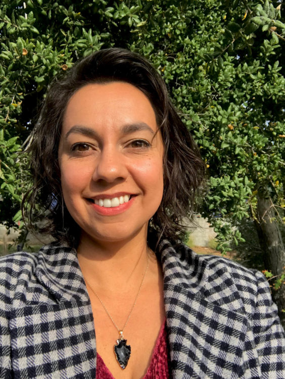 a woman in a checkered coat with wavy hair brown hair smiles toward the viewer in front of a California oak tree