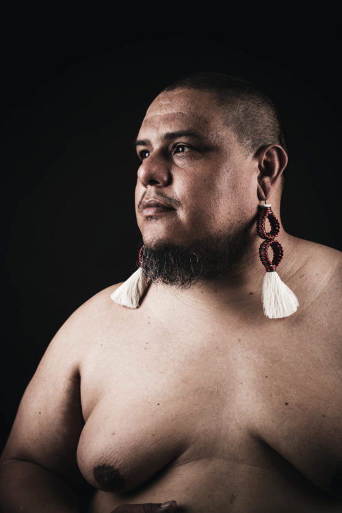 A bare-chested brown creature with a shaved head, wearing long white tassel earrings while radiating severe beauty and staring serenely to their right