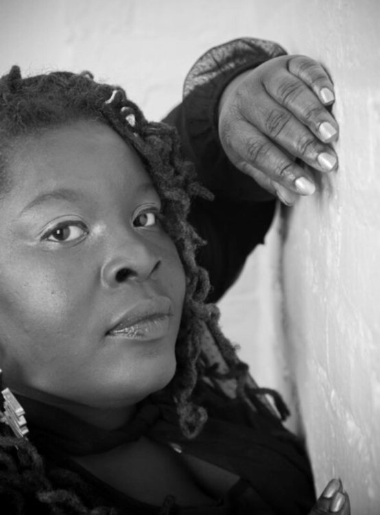 Black and white semi-profile image of a woman with back length Locs looking seriously at the viewer, with one hand propped on a white brick wall. Her nails are polished gold.