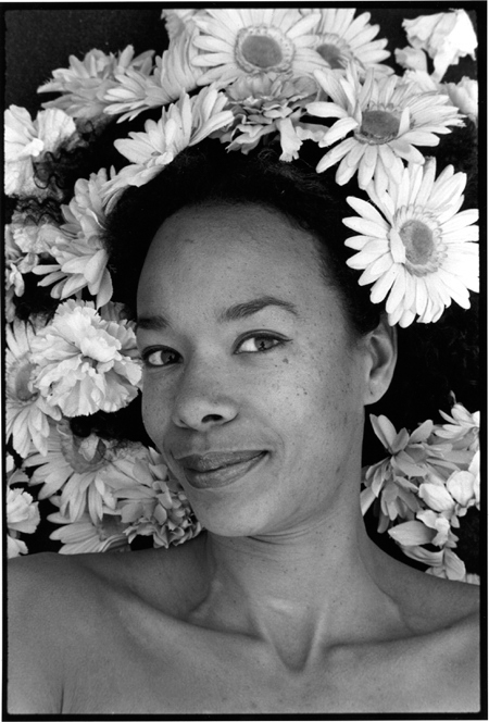 A woman of African descent smiling into the camera. Her hair is festooned with carnations and gerbera daisy flowers, that surround her face like a halo.