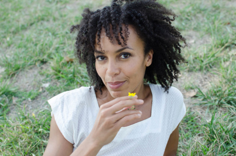 A black woman with curls tumbling in the wind has just smelled a delicate yellow flower and reveals an inner pleasure from the scent
