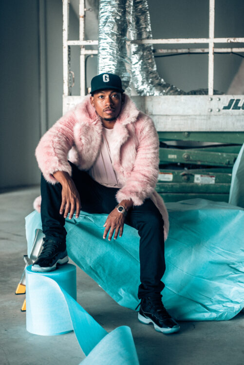 A Black man with a baseball hat on his head, wearing a pink fur coat and pink teeshirt. He is sitting on a ladder with his feet on a roll of foam. Behind him is a commercial hydraulic lift.