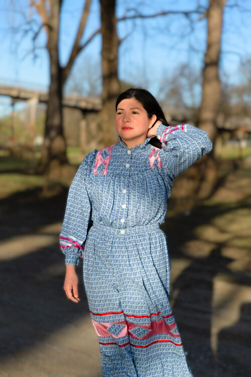 An Indigineous woman with dark hair is wearing a blue and pink Cherokee tear dress.