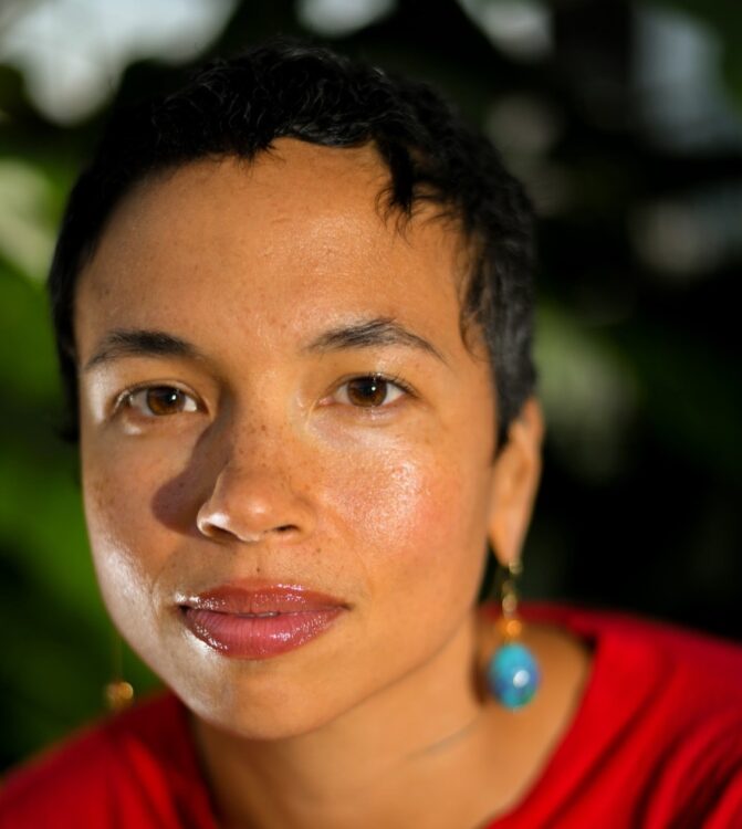 Headshot of artist and performer, Holland Andrews who has short brown hair, light brown skin, brown eyes, wearing a red shirt and looking into the camera with a black background.