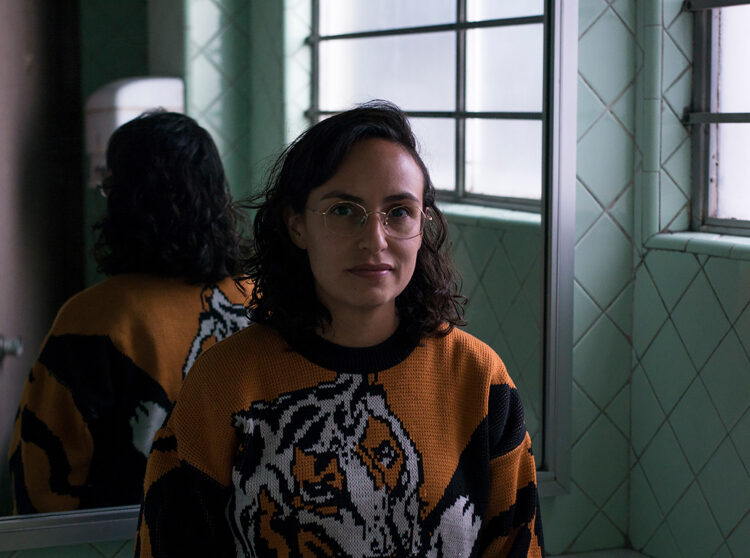 A brown skin woman with short wavy dark hair, wears a knitted sweater with an orange and black tiger figure, stands with her back to a mirror that reflects the back of her head and tiger sweater. Around the mirror, there are mint blue tiles and an opaque window.