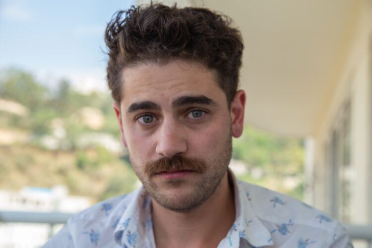 Mustached man staring above the camera. He has a white collared shirt with blue floral designs, and is looking into the lens. He is outside but the background is out of focus.