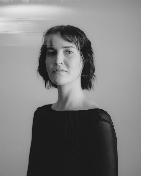 A non-binary individual with shoulder length hair, wearing all black in a empty space.