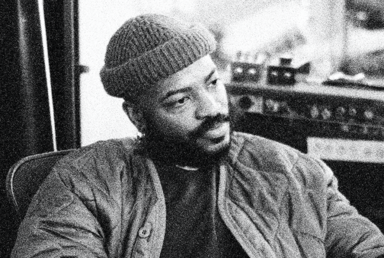 A black and white photograph of a bearded black man seated in a chair inside a recording studio, gazing off into the distance, wearing a beanie.