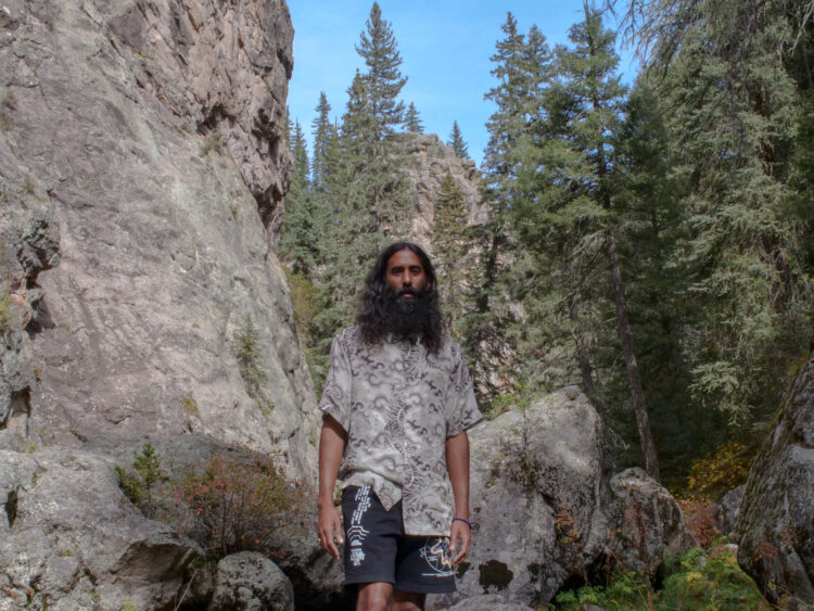 A brown man with black shoulder length hair and a beard stands in a grey stone canyon filled with pine trees under a blue sky. He wears a silver and black patterned shirt and black printed shorts.