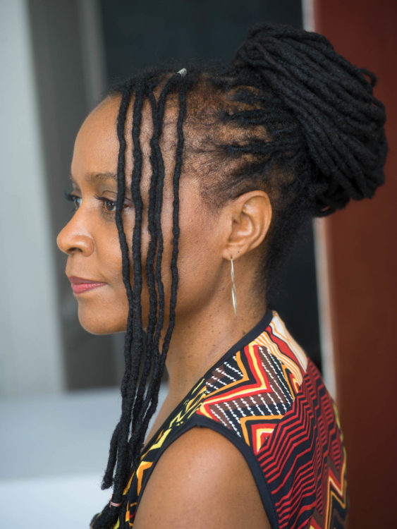 A woman in profile with dread locs piled up on her head, smiles quietly to herself.