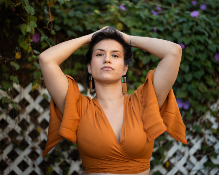 A light brown woman is looking at the camera with her chin up and her hands in her hair, wearing turquoise earrings and an orange top. Behind her is an overgrown hedge with purple flowers and a white fence.