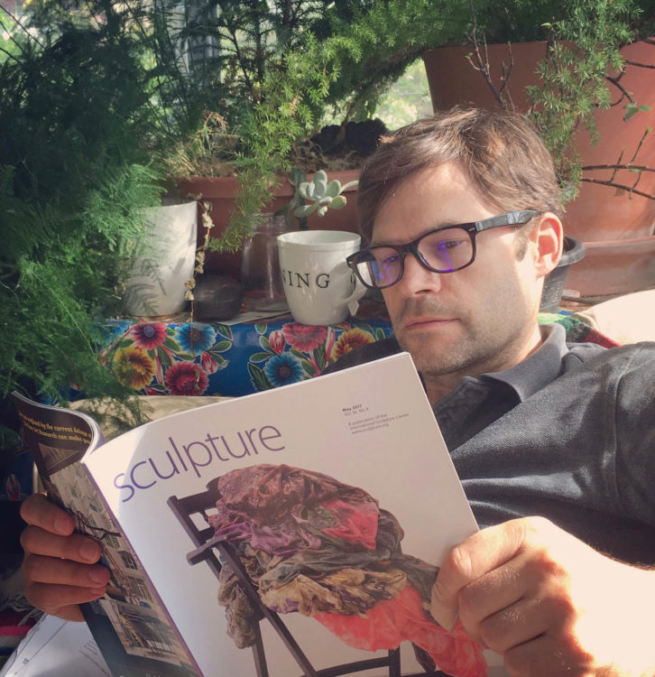 A man wearing glasses reads Sculpture magazine in a light filled sunroom surrounded by plants.