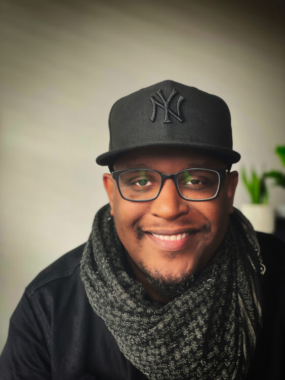 A black man smiling, wearing black rimmed glasses with an off-white background in a black Yankees hat and a black and grey scarf.