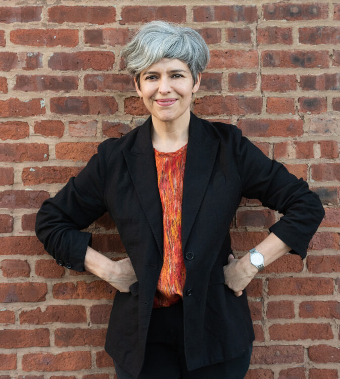 Mónica de la Torre's headshot. A silver-haired olive-skinned woman with a short, asymmetrical haircut wearing a red patterned blouse and a black blazer. Her hands rest on her hips and she leans against a brick wall.