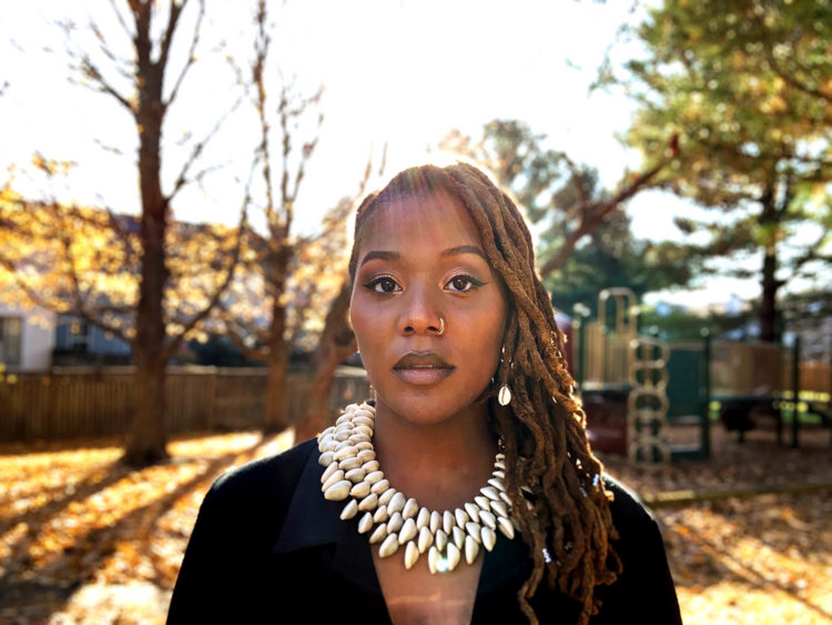 A black woman with shoulder length locs and cowrie shell necklace looks towards the viewer emanating stoicism.