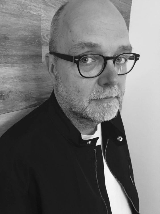 A bearded man with glasses and a black jacket looks pensively at the photographer.