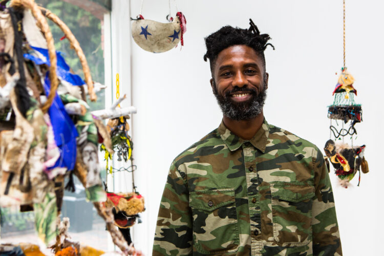 A Black man with full beard and mustache, wearing a camouflage shirt smiles slightly. He is surrounded by small floating, tactile vessels made of various materials. There is a white wall and window behind the person and vessels. Photo by Melissa Blackall