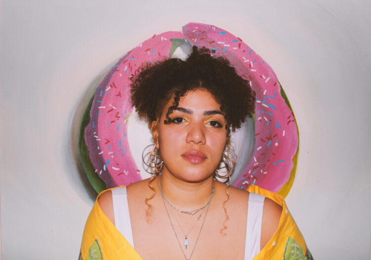 A woman with curly hair up in a pony tails, wearing a yellow off the shoulder shirt and matching eyeshadow sits framed by a painting. The painting The painting behind her is comprised of two plantains making the shape of a donut, frosted with pink icing and red, white and blue sprinkles.